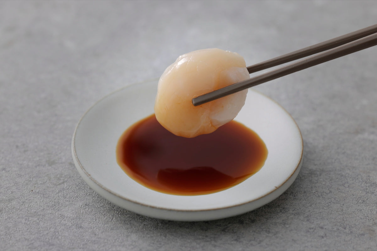 箸でつまんだ帆立のお刺身を醤油につけるところ